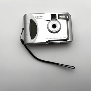 May include: Silver digital DSC Pro camera with a black wrist strap. The camera has a lens, flash, and the text "True Digital VGA 640x480 - 2MB". The camera is on a white surface.
