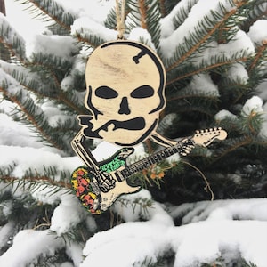May include: A wooden skull ornament playing a guitar with a colorful design. The ornament is hanging on a snowy evergreen tree branch.