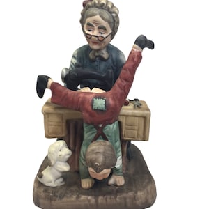 May include: A porcelain figurine of a grandmother sewing while a child does a handstand over the sewing machine. A small white dog is watching the scene.