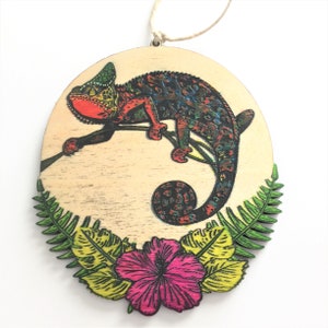 May include: A wooden ornament shaped like an oval with a colorful chameleon on a branch with green leaves and a pink flower. The chameleon is red, green, blue, and black.