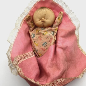 May include: A vintage baby doll with a round face and short brown hair, wrapped in a pink blanket with a lace trim. The doll is wearing a patterned onesie with animal figures. The blanket is nestled in a white lace-trimmed cradle.