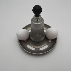 May include: A vintage egg separator made of metal, with a black handle and a circular base. Two white eggs sit on the base. The separator is designed to separate egg whites from yolks. The background is white.