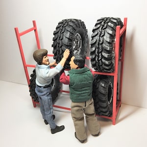 May include: Two people are standing next to a red metal rack holding four black tires. The tires have a tread pattern and are stacked on top of each other.