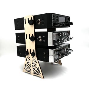 May include: A wooden stand with a tower design holds three black CB radios. The radios have silver accents and knobs. The stand is made of light wood and has a geometric pattern.