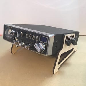 May include: A black CB radio with a silver faceplate and a wooden stand. The radio has a digital display and several knobs and buttons. The text "STRYKER" is visible on the radio.