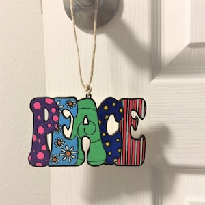 May include: A wooden peace sign ornament with a colorful, psychedelic design. The sign is hanging from a string and has a pink, blue, green, and red color scheme.