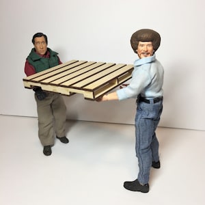 May include: Two figures are holding a wooden pallet. The pallet is made of light wood with brown edges. The figures are wearing casual clothing.