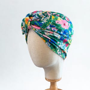 Może przedstawiać: Kwiatowy turban w odcieniach zieleni, różu, błękitu i żółci, prezentowany na głowie manekina. Materiał jest marszczony i zawiązany u góry, tworząc stylowe nakrycie głowy. Turban to żywy dodatek.