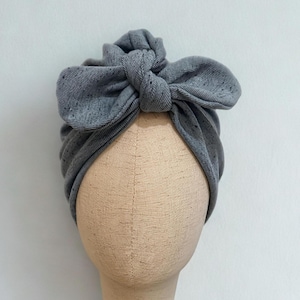 Könnte beinhalten: Ein graues Turban-Stirnband mit einer großen Schleife oben, präsentiert auf einem Schaufensterpuppenkopf. Das Stirnband ist aus einem weichen, strukturierten Stoff gefertigt. Der Schaufensterpuppenkopf ist beige und auf einem Holzständer montiert.