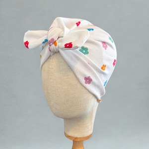 May include: A white turban-style head wrap with a tied bow, adorned with a pattern of small, colorful flowers. The flowers are in shades of red, blue, orange, green, and pink. The head wrap is displayed on a mannequin head.
