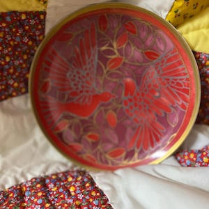 May include: A round decorative plate with a bird design. The plate features a red and pink bird and leaf design with gold accents. The plate has a gold rim and is set against a patterned background.