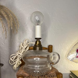 May include: A clear glass oil lamp with a brass base and a lightbulb. The lamp is shaped like a teapot and sits on a small wooden chair with a woven seat. A white electrical cord is coiled nearby.
