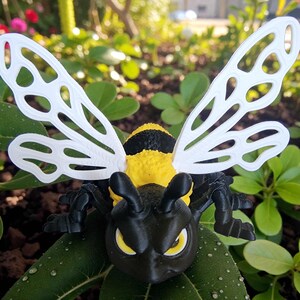 May include: A black, yellow, and white bee figurine with detailed wings and a stern expression. The bee has a black head and legs, a yellow and black striped body, and white wings. The figurine is set against a backdrop of green leaves.