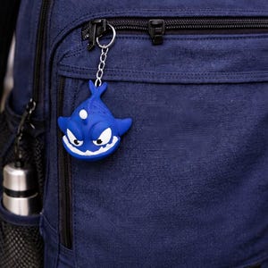 May include: A blue backpack with a shark keychain. The keychain is a cartoon shark with white accents and a chain. The backpack is a dark blue color with black zippers and straps. A water bottle is in the side pocket.