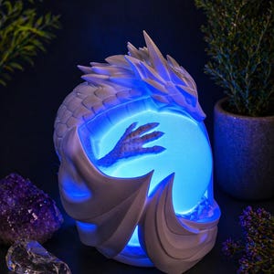 May include: A white dragon sculpture holds a glowing blue orb. The dragon's detailed scales and wings contrast with the smooth, illuminated sphere. The sculpture is a fantasy-themed decorative item.