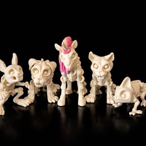 May include: Five off-white skeleton figurines of a rabbit, tiger, unicorn, cat, and lizard. The unicorn has a pink mane and horn. The figurines are displayed on a black surface.