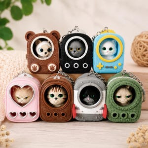 May include: A collection of eight cat-themed keychains. Each keychain features a different cat inside a unique, colorful casing. Designs include a bear, a game console, a rocket, and a heart. The keychains are made of a hard material and have silver-colored chains.