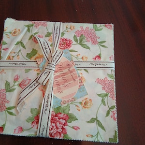 May include: A square stack of floral fabric squares tied with a ribbon. The fabric features a cream background with pink, red, and yellow flowers and green leaves. A tag with the words "Nature's Chorus" is attached.