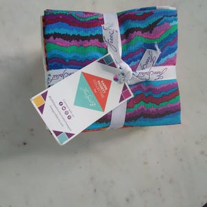 May include: A stack of colorful fabric squares tied with a white ribbon. The fabric features a wavy pattern in shades of blue, purple, green, and pink. A tag with the text "KAFFE FASSETT COLLECTIVE for Free Spirit" is attached.