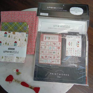 May include: A collection of Christmas-themed craft supplies, including fabric bundles with red and green patterns, a countdown panel, and embroidery floss. The fabric bundles are tied with a white ribbon that reads "snowkissed".