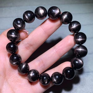 Puede incluir: Una pulsera compuesta por cuentas de obsidiana negra pulidas, lisas y redondas. Cada cuenta presenta un brillo plateado distintivo y reflectante que crea un efecto metálico brillante en la superficie de las piedras oscuras y circulares.