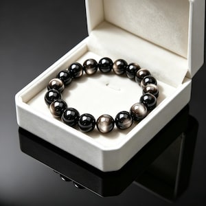 May include: A circular bracelet composed of polished, spherical black and silver sheen obsidian beads. The beads exhibit a reflective, metallic lustre with alternating dark and light grey tones. The bracelet is displayed inside an open white box.