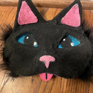 May include: A black cat mask with blue mesh eyes, pink inner ears, and a pink nose and tongue. The mask has black fur around the edges and is likely for a costume or theatrical use.