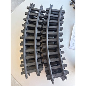 Puede incluir: Vías de tren curvas de plástico gris oscuro. Las vías están diseñadas para conectarse y formar un diseño circular u ovalado. La imagen muestra varios juegos de vías, listos para un juego de trenes modelo.
