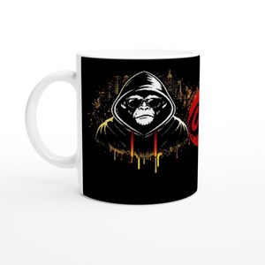Könnte beinhalten: Weiße Keramik-Tasse mit einer Grafik eines Schimpansen mit Kapuzenpullover und Sonnenbrille, vor einer Stadtkulisse. Das Design enthält rote und goldene Akzente, mit dem Wort "OG" in Rot.