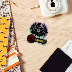 May include: A holographic sticker with a gorilla in a hoodie and sunglasses, and the text "City Monkey". The sticker has a cityscape silhouette and a red and green circular design. The sticker is on a wooden surface with travel documents and a camera.