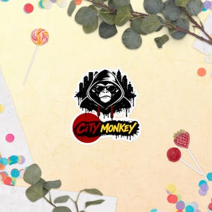 Könnte beinhalten: Ein Aufkleber mit einer schwarz-weißen Grafik eines Affen mit Kapuzenpullover und Sonnenbrille, vor einer Skyline-Silhouette. Der Text "City Monkey" ist in einer stilisierten Schriftart dargestellt.
