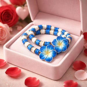 Puede incluir: Dos pulseras con cuentas azules, blancas y doradas, adornadas con detalles florales azules y amarillos, descansan dentro de una caja de joyería rosa. Las pulseras están rodeadas de pétalos de rosa y rosas, creando un tema romántico.