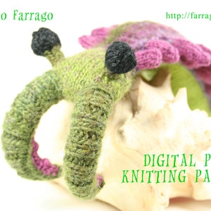 May include: A green and purple knitted amigurumi creature with black eyes. The creature is sitting on a white seashell. The text "DIGITAL PDF KNITTING PATTERN" is written in green letters on the seashell.