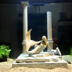 May include: A detailed, off-white mermaid figurine with flowing hair, reclining on a stone platform. Two classical columns frame the mermaid, with a broken column fragment nearby. The scene evokes an ancient, aquatic theme.
