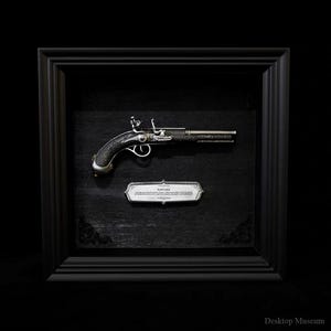 May include: A framed antique flintlock pistol, presented on a black background. The pistol has detailed silver embellishments on the handle and barrel. A small silver plaque below the pistol has text. The frame is black.