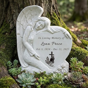 Könnte beinhalten: Eine weiße Engelsstatue mit ausgebreiteten Flügeln, die sich an einem herzförmigen Denkmal abstützt. Auf dem Denkmal steht "In Loving Memory of Lena Proce" mit Daten. Die Statue ist von Grün und Sukkulenten umgeben.