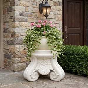 May include: A white, ornate plant stand with a large, round planter filled with pink flowers and trailing green and white foliage. The stand has a decorative base with scrollwork and is placed outside a home.