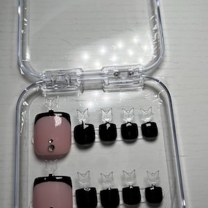 May include: A set of artificial nails in a clear plastic case. The nails feature a pink base with a black French tip and small rhinestones. The case is open, displaying the nails in two rows.