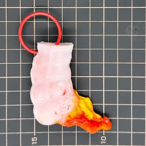 Puede incluir: Un llavero hecho a mano, de forma abstracta, con un anillo rojo. El cuerpo principal es blanco con acentos rosas y rojos, que transicionan a amarillo y naranja en la base. El llavero está sobre un fondo de cuadrícula.