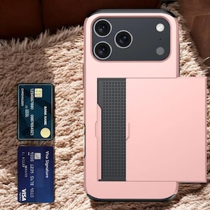 May include: A pink phone case with a black camera module and a cardholder slot. Two credit cards are next to the phone case. One is blue with the Visa logo, and the other is dark blue with the text "CARDHOLDER".