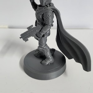May include: A gray 3D printed miniature figure of a soldier wearing a helmet and armor with a skull emblem on the shoulder. The figure holds a rifle in one hand, has a raised fist, and wears a long flowing cape, all standing on a circular base.