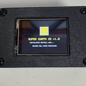 May include: A black rectangular electronic device with a screen displaying a yellow skull icon and the text SUPER EARTH OS v1.0, INITIALIZING TACTICAL LINK..., and WELCOME HELL DIVER MISSILDINE. The screen also shows the text TPM408-2.8 on the left side.