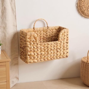 May include: A rectangular, woven basket with a handle, mounted on a wall. The basket is made of natural-colored fibers and has a textured appearance. The basket is designed for storage and organization.