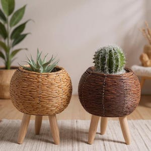 Op de afbeelding: Twee ronde plantenbakken van geweven riet met houten poten. Eén is lichtbruin en bevat een vetplant, de andere is donkerbruin met een cactus. De plantenbakken hebben een natuurlijke, rustieke uitstraling.