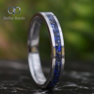Könnte beinhalten: Ein silberner Ring mit einem Band aus zerkleinerten blauen und goldenen Steinen. Der Ring hat eine polierte Oberfläche und die Worte "Stellar Bands" befinden sich in der oberen linken Ecke. Der Hintergrund ist ein verschwommenes Grün.