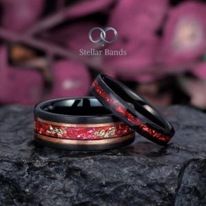 May include: Two black rings with red and gold accents. One ring features a band of red and gold embedded in a wood-like material, while the other has a red inlay. The rings are displayed on a dark, textured surface.