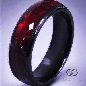 Puede incluir: Un anillo negro con una banda de piedras preciosas facetadas de color rojo intenso. El anillo tiene un acabado brillante y un perfil redondeado. El logotipo de Stellar Bands es visible en la esquina inferior derecha.