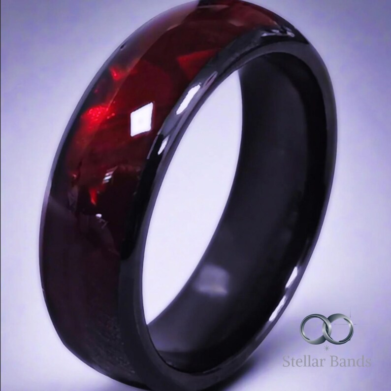Puede incluir: Un anillo negro con una banda de piedras preciosas facetadas de color rojo intenso. El anillo tiene un acabado brillante y un perfil redondeado. El logotipo de Stellar Bands es visible en la esquina inferior derecha.