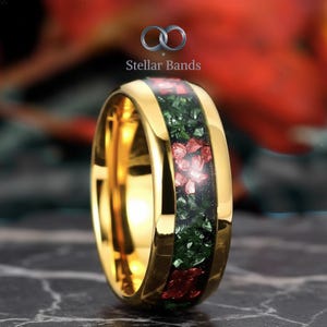 May include: A gold-colored ring with a central band of green and red crushed stone. The ring has a polished finish and the text "Stellar Bands" is visible at the top.
