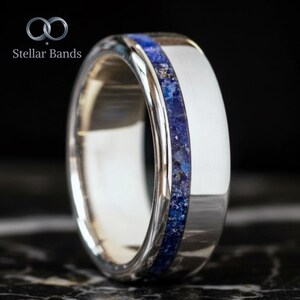 May include: A silver ring with a wide band and a central inlay of deep blue lapis lazuli. The ring has a polished finish and the text "Stellar Bands" is visible in the upper left corner.